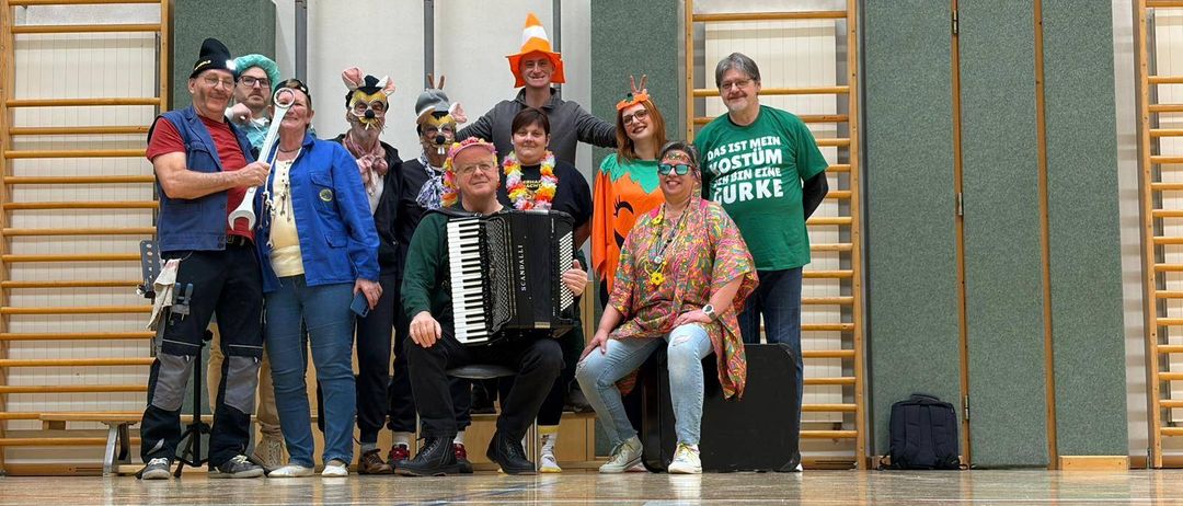 Eine Gruppe von Menschen in Kostümen steht und sitzt auf einem Turnhallenboden. Ein Mann spielt Akkordeon.