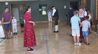 Eine Gruppe von Menschen befindet sich in einem Raum mit Holzboden. Eine Frau in einem roten Kleid steht vorne, und ein Mann in einem Weste steht hinter ihr. Eine Frau in einem blauen Shirt macht ein Foto.
