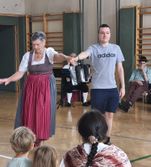 Eine ältere Frau in traditioneller Kleidung spielt Akkordeon, während ein Mann in lässiger Kleidung neben ihr steht, und ein sitzender Mann spielt Klavier im Hintergrund, während mehrere Kinder davor zusehen.