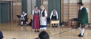 Eine Gruppe von Menschen in traditioneller Kleidung führt auf einer Bühne auf. Zwei Frauen stehen vorne, eine spielt Akkordeon, die andere singt. Hinter ihnen spielt ein Mann Keyboard. Das Publikum sitzt vor ihnen.
