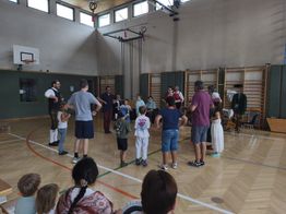 Eine Gruppe von Menschen, einschließlich Kindern, versammelt sich in einer Turnhalle. Einige stehen im Kreis, während andere von den Seitenlinien zuschauen. Im Hintergrund sind Geräte und Instrumente zu sehen.