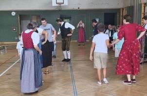 Eine Gruppe von Menschen in traditioneller Kleidung tanzt in einer Turnhalle. Ein Mann spielt Akkordeon, während andere zusehen. Ein Basketballkorb ist im Hintergrund zu sehen.