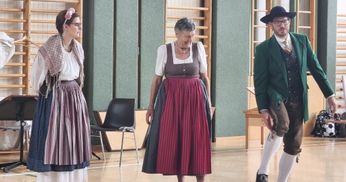 Eine ältere Frau in einem traditionellen Kleid mit einem roten Rock und einem weißen Oberteil steht in einem Raum mit einem Holzstuhl und Holzregalen im Hintergrund.