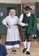 Ein Mann und eine Frau in traditioneller deutscher Kleidung tanzen gemeinsam in einem Raum. Die Frau trägt ein weißes Kleid und einen Hut, während der Mann eine Weste, eine Krawatte und braune Hosen trägt. Ein anderer Mann spielt im Hintergrund Akkordeon.