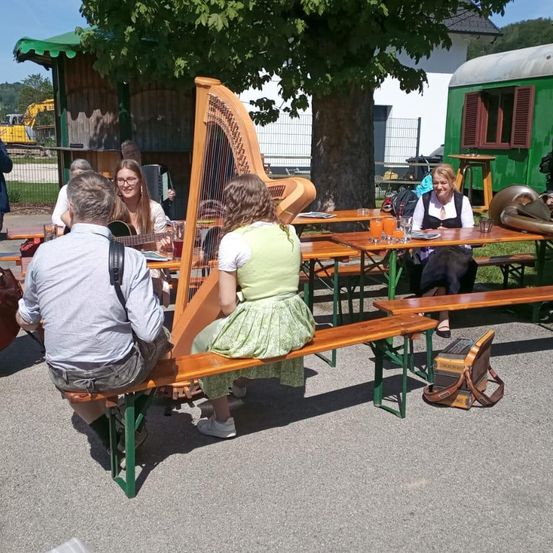 Eine Gruppe von Menschen sitzt auf Holzbänken, einige spielen Instrumente. Eine Frau spielt Harfe, eine andere Gitarre. Eine Frau sitzt allein mit einer Tasche auf dem Schoß. Ein Baum ist im Hintergrund.