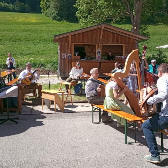 Eine Gruppe von Musikern spielt auf Bänken vor einem Holzschuppen, wobei eine Person die Harfe und eine andere das Cello spielt. Eine Frau sitzt vor einem Tisch, und eine Person steht neben ihr. Hinter ihnen stehen mehrere Personen.