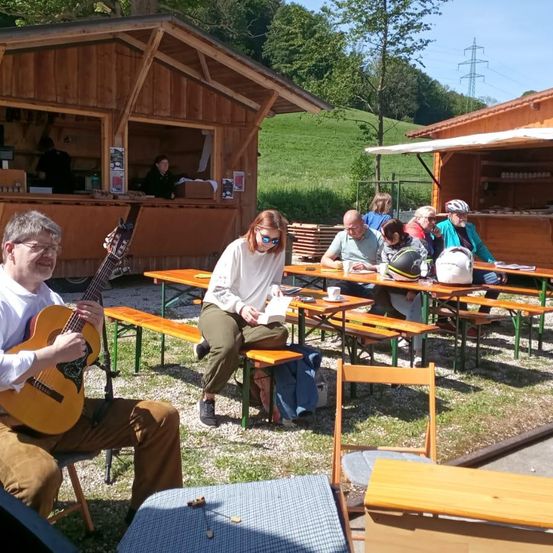 Eine Gruppe von Menschen sitzt um einen Holztisch im Freien. Ein Mann spielt Gitarre. Andere Menschen sind in einem kleinen Holzgebäude.
