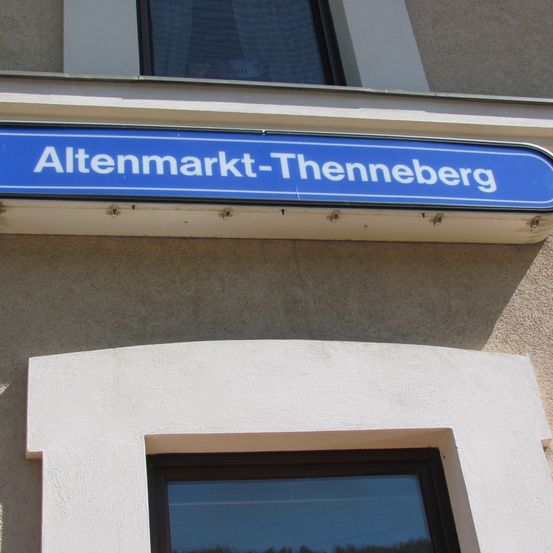 Ein blaues Schild mit weißen Buchstaben, das 'Altenmarkt-Thenneberg' an der Außenwand eines Gebäudes befestigt ist.