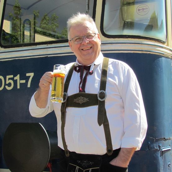 Ein Mann mit Brille hält ein Glas Bier vor einem blauen Zug. Er trägt ein weißes Hemd, Hosenträger und eine Fliege.
