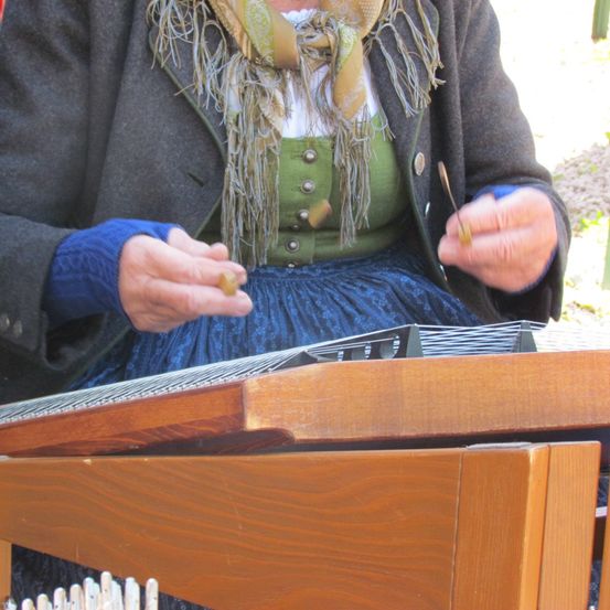 Eine Person in einem grünen Weste und einem Schal spielt ein Instrument aus Holz mit beiden Händen.
