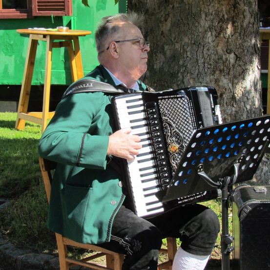 Ein älterer Mann in einem grünen Mantel und einer Brille spielt ein Akkordeon mit einem schwarzen Ständer in einem Park unter einem Baum.