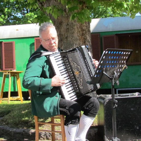 Ein älterer Mann in einer grünen Jacke und weißen Socken spielt ein Akkordeon unter einem Baum in der Nähe eines grünen Zugwaggons.