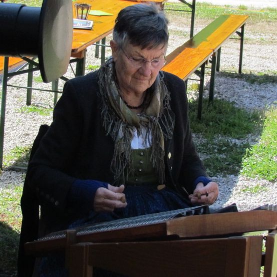 Eine ältere Frau mit Brille und Schal sitzt und spielt Hackbrett, mit Bänken und einem Tisch dahinter.