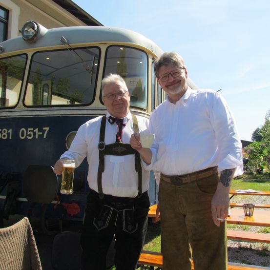Zwei Männer stehen neben einem Zug, einer hält ein Glas Bier. Sie tragen traditionelle deutsche Kleidung. Der Zug zeigt eine Nummer an.