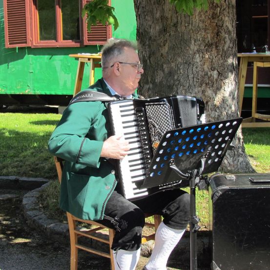 Ein älterer Mann in grüner traditioneller Kleidung spielt Akkordeon, auf einem Stuhl unter einem Baum sitzend, mit einem Notenständer vor ihm.