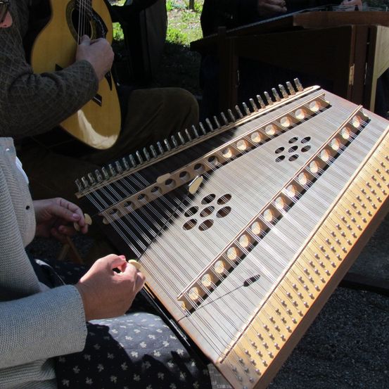 Eine Person spielt eine Zither, während sie auf dem Boden sitzt. Eine weitere Person hinter ihr spielt eine Gitarre. Ein Tisch steht neben ihnen.