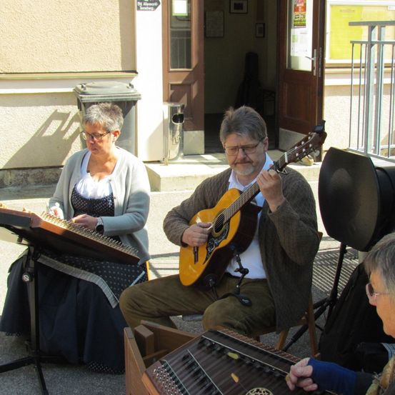 Zwei Erwachsene sitzen auf dem Bürgersteig und spielen Musikinstrumente. Der Mann spielt Gitarre, die Frau Zither. Ein Lautsprecher und ein Mikrofon sind in der Nähe.