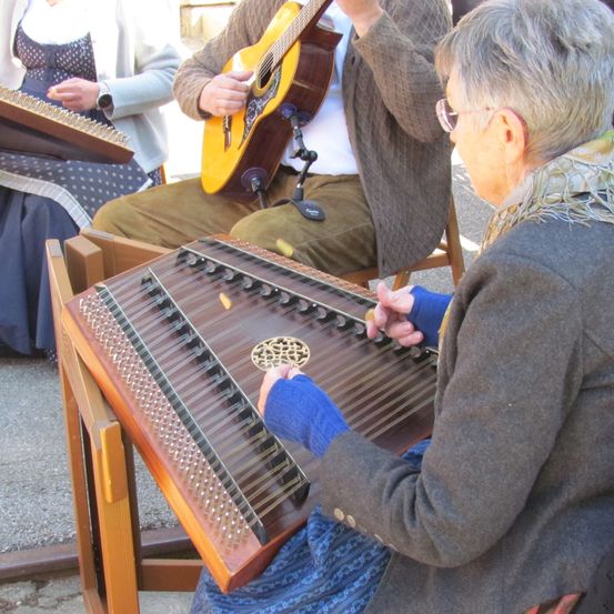 Eine ältere Frau spielt eine Zither, während sie sitzt, mit einem Gitarristen hinter ihr und einer weiteren Person mit einer Zither auf der linken Seite. Sie sind draußen, wahrscheinlich bei einer Aufführung.