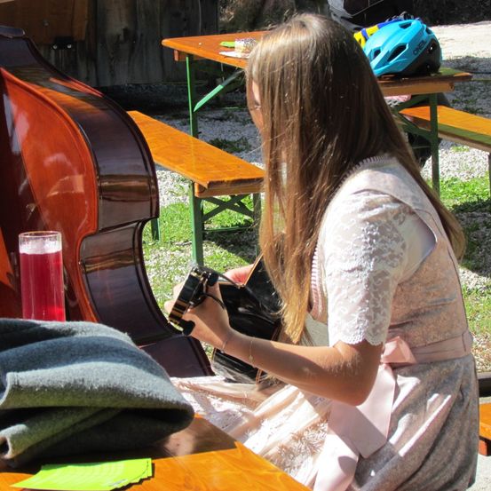 Ein Mädchen sitzt draußen auf einer Bank und spielt Cello. Sie trägt ein rosa Kleid. Ein Glas mit roter Flüssigkeit und ein Helm stehen in der Nähe.