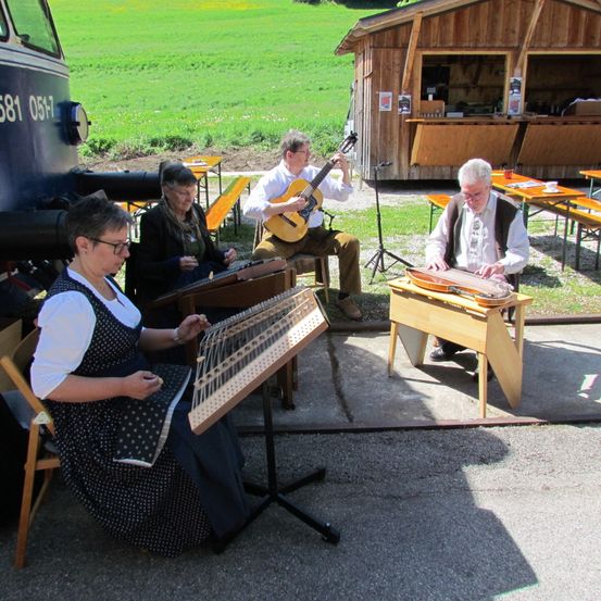 Vier Musiker, zwei Männer und zwei Frauen, spielen traditionelle Instrumente. Ein Mann spielt Gitarre, der andere Zither. Eine Frau spielt Zither, und eine andere Frau spielt Hackbrett. Sie sitzen auf Stühlen mit einer kleinen Bühne davor. Dahinter steht ein kleines Holzhaus.