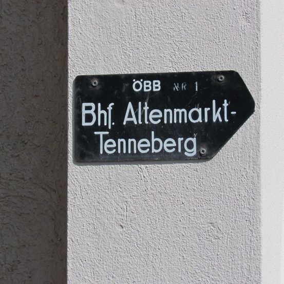 Ein schwarzes Schild mit weißen Buchstaben an einer weißen Wand weist den Weg zum Bahnhof Altenmarkt-Zennerberg.