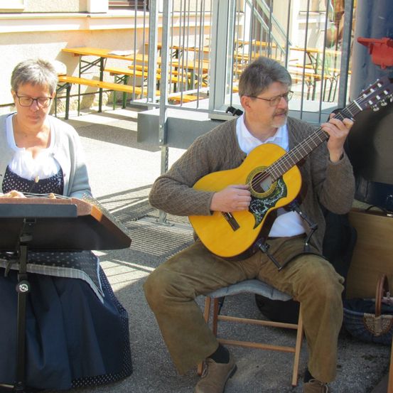 Ein Mann mit Brille und einem braunen Schal spielt eine gelbe akustische Gitarre, während er auf einem Stuhl sitzt. Eine Frau mit Brille und einem blauen Kleid liest Noten auf einem Notenständer. Dahinter steht ein Metallzaun mit Holzbänken.