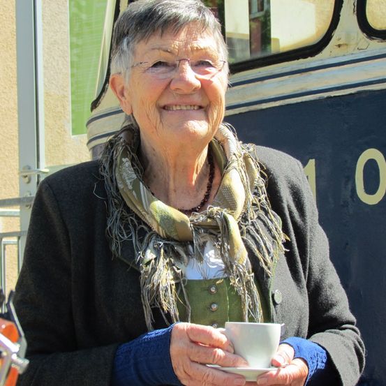 Eine ältere Frau lächelt, während sie eine weiße Tasse in den Händen hält. Sie trägt eine Schürze und eine Brille und steht vor einem Bus.