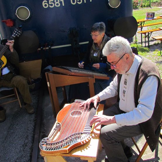 Ein älterer Mann spielt ein Zither, während ein anderer eine Gitarre vor einem Zug spielt. Eine Frau sitzt hinter ihnen. Es gibt Bänke und Tische hinter dem Zug.