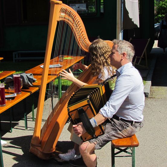 Ein erwachsener Mann und ein Mädchen spielen draußen auf einer Bank Harfe und Akkordeon. Es gibt Holzbänke mit Getränken darauf. Eine Frau geht im Hintergrund.
