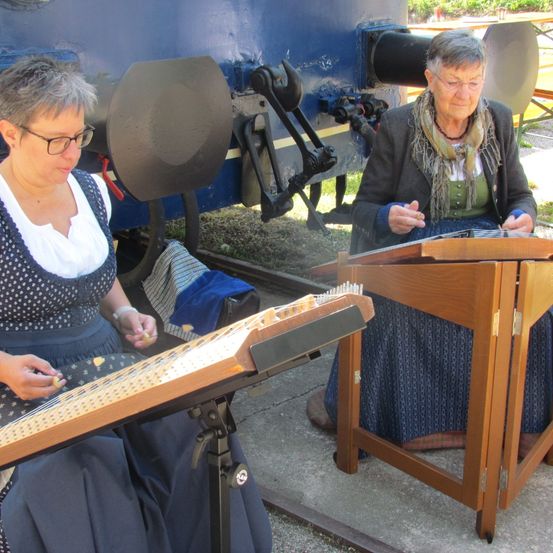 Zwei Frauen sitzen auf dem Boden und spielen jeweils ein Hammerklavier. Eine Frau trägt eine gepunktete Schürze, und sie tragen beide eine Brille. Ein Zug ist hinter ihnen.