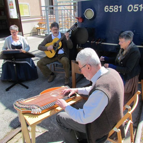 Vier ältere Personen sitzen draußen und spielen jeweils ein anderes Musikinstrument. Der Mann im Vordergrund spielt eine Harfe, während der Mann neben ihm eine Gitarre spielt. Eine Frau liest Noten auf einem Ständer. Dahinter steht ein blauer Zug mit der Nummer 6581 051.