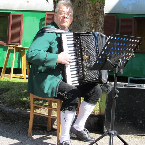 Ein älterer Mann spielt Akkordeon, während er auf einem Holzstuhl sitzt. Er ist in einer grünen Jacke und weißen Socken gekleidet. Hinter ihm steht ein grünes Gebäude mit geschlossenen Fensterläden und einem Lautsprecher.