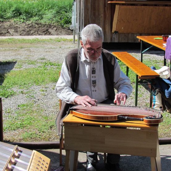 Ein älterer Mann mit Brille spielt draußen an einem Holztisch eine Zither.