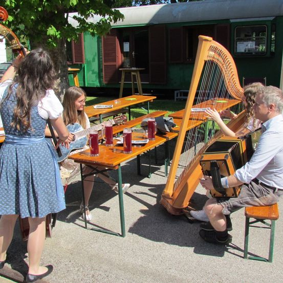 Zwei Personen spielen Harfe und Akkordeon, wobei ein Mädchen die Gitarre spielt. Sie sitzen um einen Tisch im Freien.