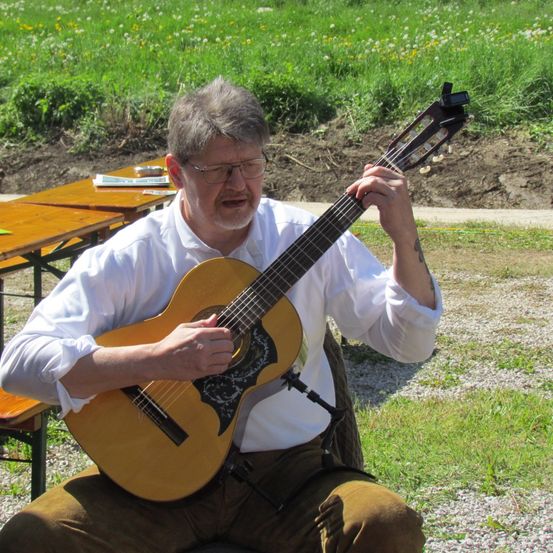 Ein älterer Mann mit Brille und weißem Hemd spielt eine Gitarre im Freien auf einer Bank mit Gras und Blumen im Hintergrund.