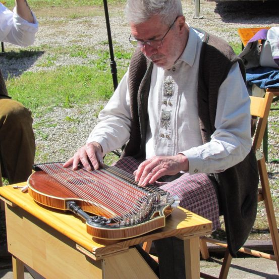 Ein älterer Mann mit Brille und Weste spielt ein Saiteninstrument auf einem Holzständer in einem Grasbereich.