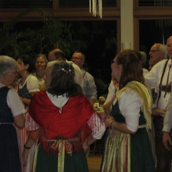 Eine Gruppe älterer Menschen ist versammelt, möglicherweise bei einer gesellschaftlichen Veranstaltung. Sie sind in traditioneller Kleidung gekleidet. Einige unterhalten sich. Im Hintergrund befinden sich eine Pflanze und eine Hängelampe.