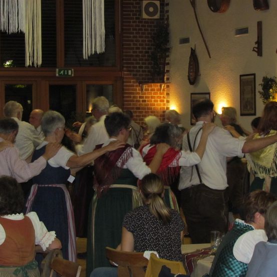 Eine Gruppe von Menschen, die in traditioneller Kleidung gekleidet sind, tanzt zusammen in einem Raum mit Ziegelwänden und gerahmten Bildern an den Wänden. Einige halten sich an den Händen.