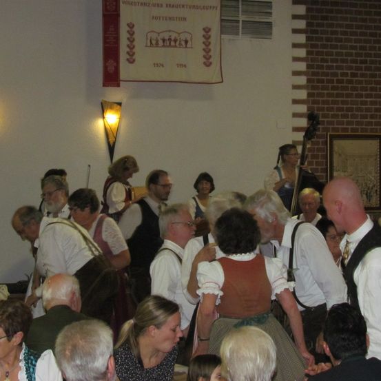 Eine geschlossene Versammlung von Menschen in traditioneller Kleidung, möglicherweise bei einem Fest. Ein Banner mit roten und weißen Designs hängt an der Wand. Einige stehen, andere sitzen, und eine Person spielt Cello.