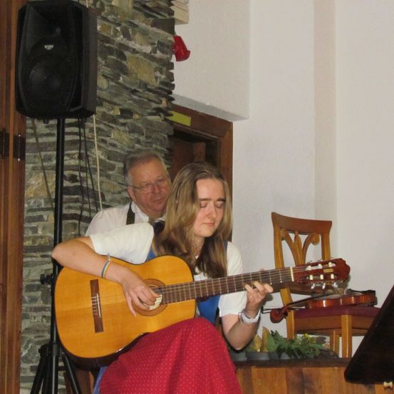 Eine Frau spielt Gitarre, während ein älterer Mann sie beobachtet. Sie befinden sich in einem Raum mit einer Steinmauer und einem Lautsprecher. Ein Holzstuhl steht in der Nähe.
