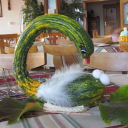 Ein aus Kürbis gefertigtes Schwan-Centerpiece mit Federn und Eiern auf einem Tisch, umgeben von Stühlen. Der Hintergrund zeigt einen Essbereich mit Pflanze und Bilderrahmen.