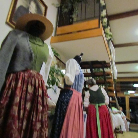 Traditionelle Kostüme auf Schaufensterpuppen in einem Raum mit Balkon und Treppe, geschmückt mit Pflanzen und Blumen. Ein gerahmtes Bild hängt an der Wand.