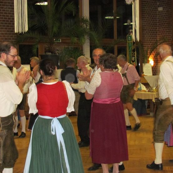 Eine Gruppe von Menschen in traditioneller deutscher Kleidung tanzt in einem Saal mit Backsteinwänden und Topfpflanzen. Einige tragen Brillen, und einer hält ein Weinglas.