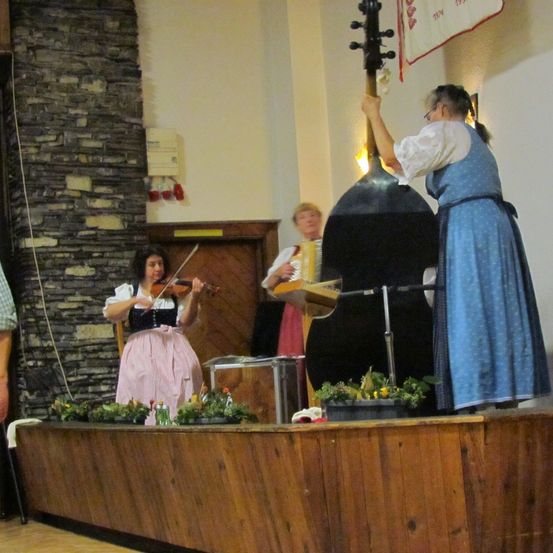 Drei Frauen auf einer Bühne spielen Musik mit einer Geige, einem Akkordeon und einer Cello. Die Bühne ist mit Blumentöpfen dekoriert. Eine Ziegelwand und eine Fahne sind im Hintergrund.