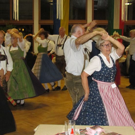 Eine Gruppe älterer Erwachsener in traditioneller Kleidung tanzt auf einem Holzboden, einige tragen Brillen. Ein Tisch mit einem Glas und einer Flasche steht im Vordergrund.