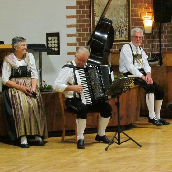 Drei Musiker spielen drinnen. Einer spielt Akkordeon, ein anderer hält ein Cello und ein Dritter sitzt auf einer Bank. Eine Frau in traditioneller Kleidung beobachtet sie.