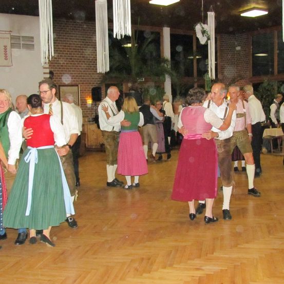 Mehrere Paare tanzen in einem Saal, einige in traditioneller Kleidung. Der Saal hat Holzböden und Ziegelwände. Im Hintergrund befinden sich Pflanzen und hängende Dekorationen.