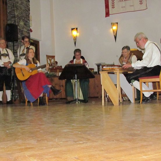 Eine Gruppe von Musikern spielt drinnen. Eine Frau spielt Gitarre, während ein Mann ein Instrument spielt. Zwei ältere Frauen beobachten, und ein Mann sitzt neben einem Tisch. Es gibt Stühle, einen Lautsprecher und eine Flagge an der Wand.