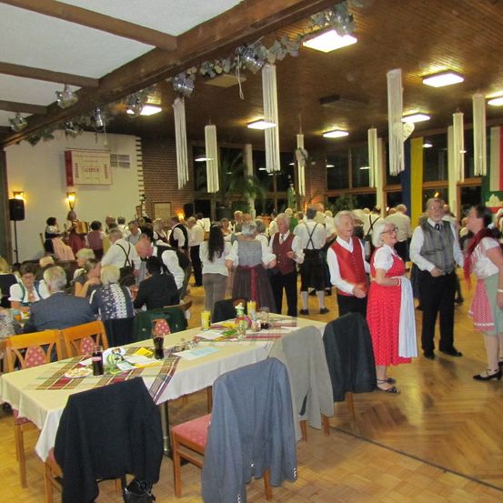 Eine festliche Veranstaltung mit traditioneller deutscher Kleidung, die Menschen im Raum sitzen und stehen, der Tisch hat Essen und Getränke, und Lichter sind an der Decke.