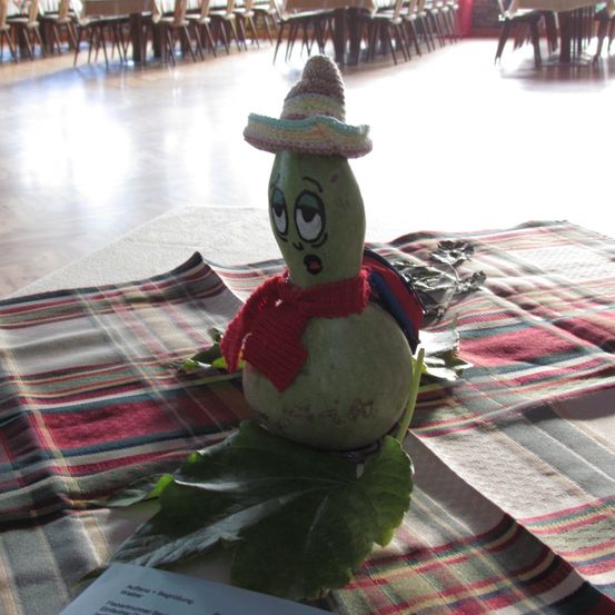 Eine Kürbisfigur mit einem Sombrero, Schal und Blättern sitzt auf einem karierten Tuch in einem Raum mit Stühlen und Tischen.